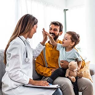 A caregiver sits on a couch with a provider, while holding a young boy in his lap. The child is holding a teddy bear and giving his medical provider a high five.