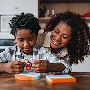 A child sits at a table playing with a push pop fidget toy while his caregiver is standing behind him smiling.