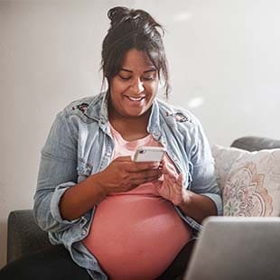 Pregnant member sitting on her couch while looking up information on her cellphone, with her laptop opened infront of her.