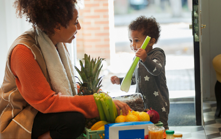 Mother carrying groceries into her home while her young son holds onto a stalk of celery.