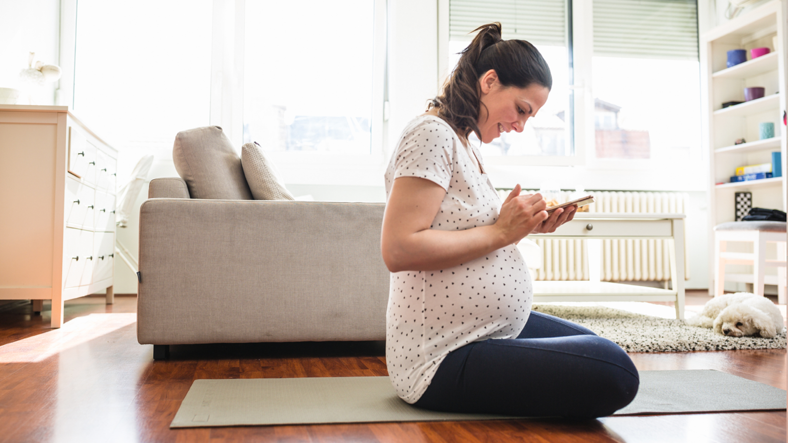 Pregnant woman sitting on the floor in living room, looking at her mobile phone.