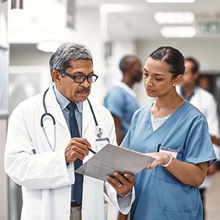 Two medical professionals stand together in a hallway reviewing their patient's medical chart.