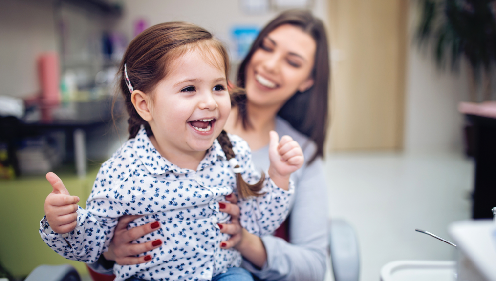 Toddler giving a thumbs up while sitting on her mother's lap at doctor's office