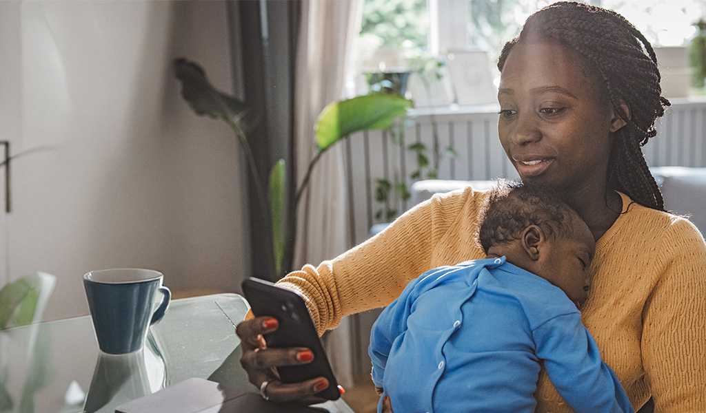 Younger mother holding her sleeping baby while looking at her phone